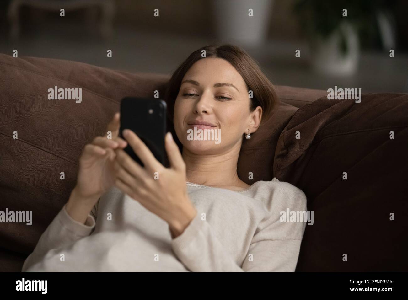Feliz mujer joven tumbada en el sofá navegar por la web en el smartphone Foto de stock