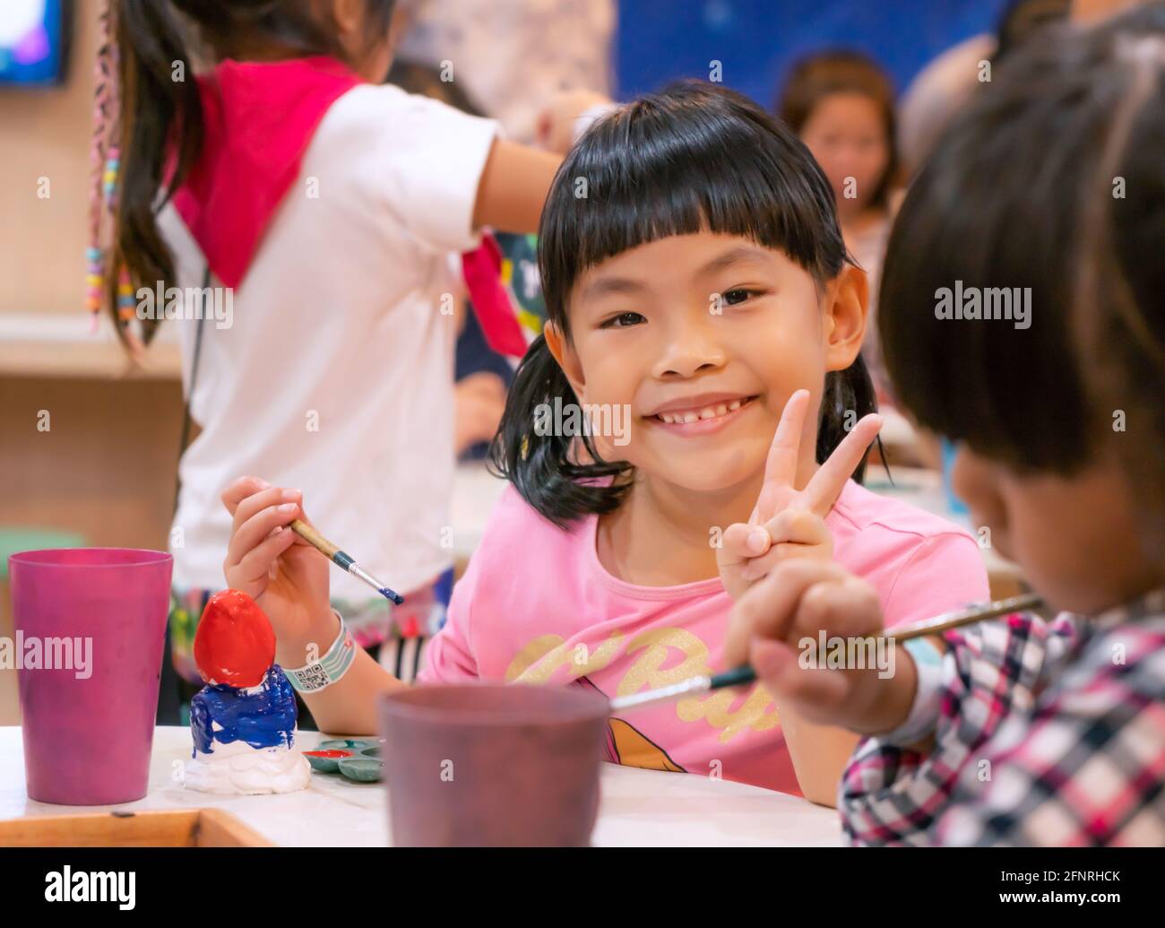 Retrato niña asiática en el taller de arte, cara feliz y sonriente, mostrar gesto dos dedos, lindo rostro con pelo negro y bang. Classmate para borroso Foto de stock