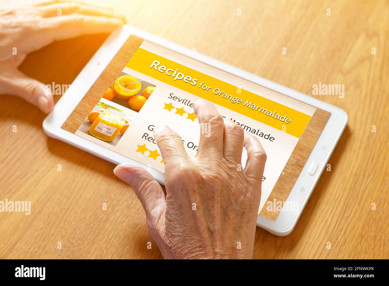 Manos de una mujer mayor tocando la pantalla de una tableta para ver una receta de mermelada de naranja, fondo de mesa de madera. Foto de stock