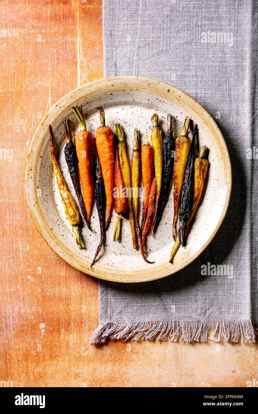 Zanahorias de diferentes colores a la parrilla en placa de cerámica con servilleta textil gris sobre fondo de piedra naranja. Vista superior, diseño plano. Espacio de copia. Cena vegana Foto de stock