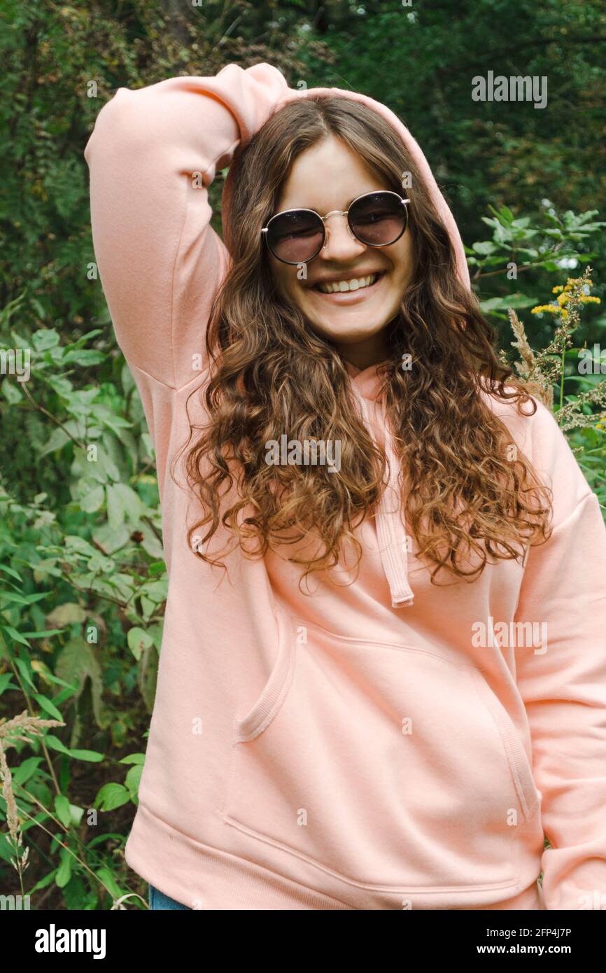 Una mujer joven con una sudadera con capucha rosa con gafas de sol, con pelo rizado, sonríe. Foto de stock