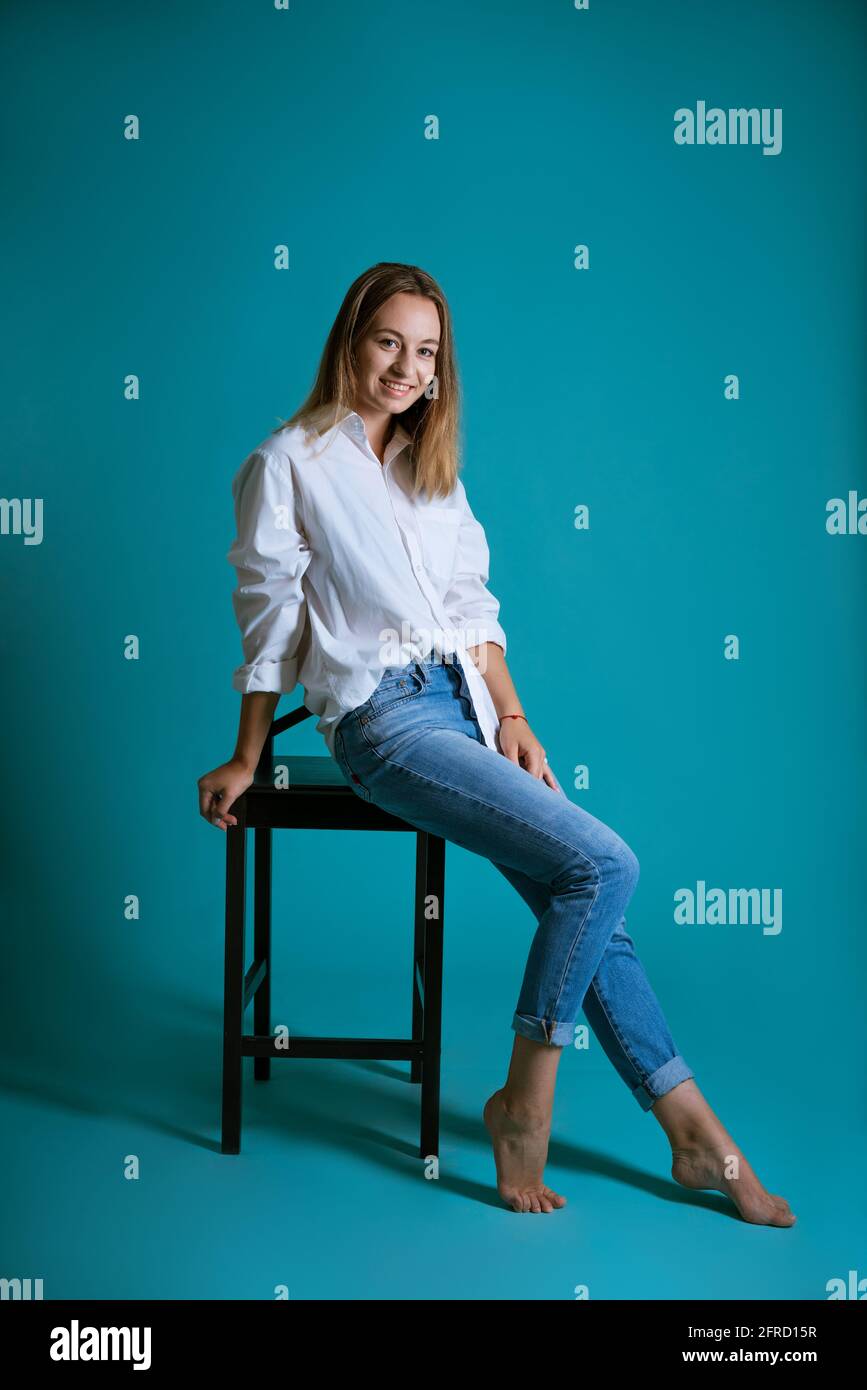 Joven hermosa mujer posando en una silla en una camisa blanca y jeans sobre un fondo azul descalzo Fotografía de stock -