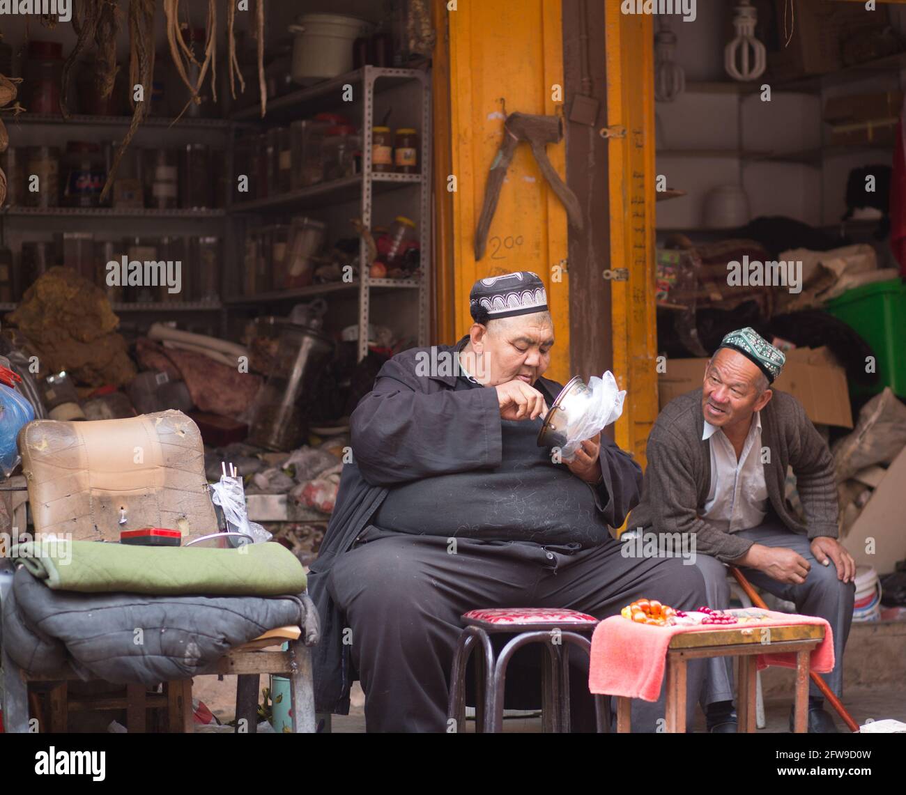 Hombre grande sentado fuera de su tienda comiendo y charlando con un amigo Kashgar, Xinkiang, República Popular China, 2019 Foto de stock