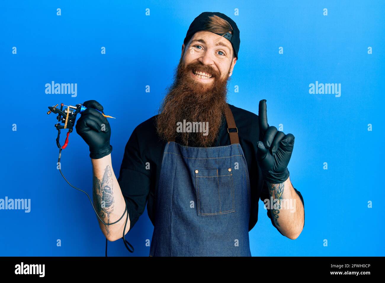 Hombre pelirrojo con un tatuador de barba larga con uniforme profesional y  guantes con una gran sonrisa en la cara, apuntando con el dedo de la mano  hacia el lado Fotografía de