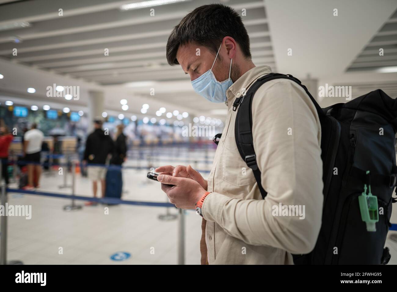 El pasajero masculino en máscara con mochila utiliza un smartphone y se  encuentra con distancia social en la línea de facturación en el aeropuerto  de Chipre, ciudad de paphos. Viaje Fotografía de