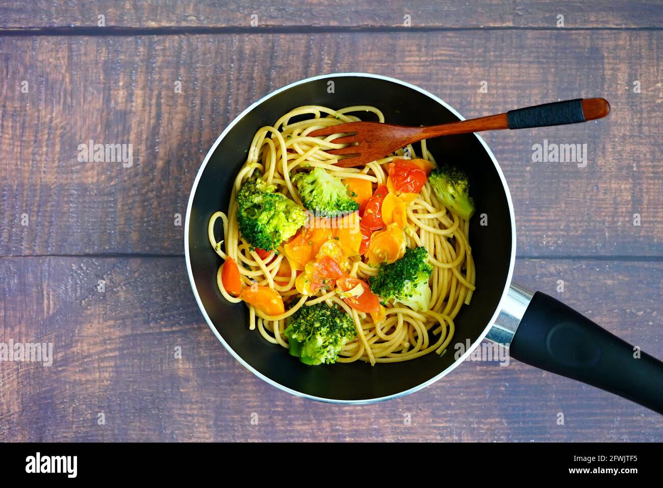 Cocina casera sana: Spagetthi con verduras (brokkoli, tomates, zanahorias) en una sartén con cuchara de madera. Foto de stock