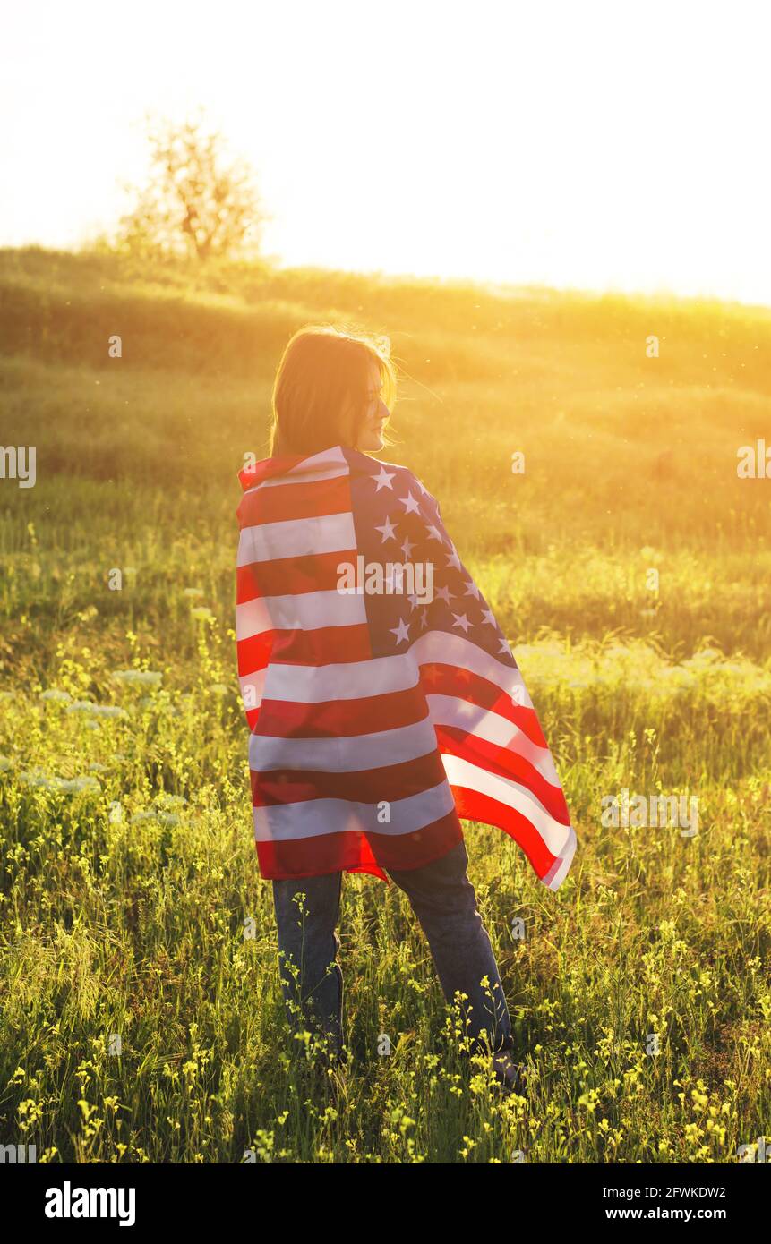 Vista posterior de una joven que sostiene la bandera nacional de los Estados Unidos en un campo al atardecer. Día de la Independencia de América. Concepto de libertad estadounidense Foto de stock