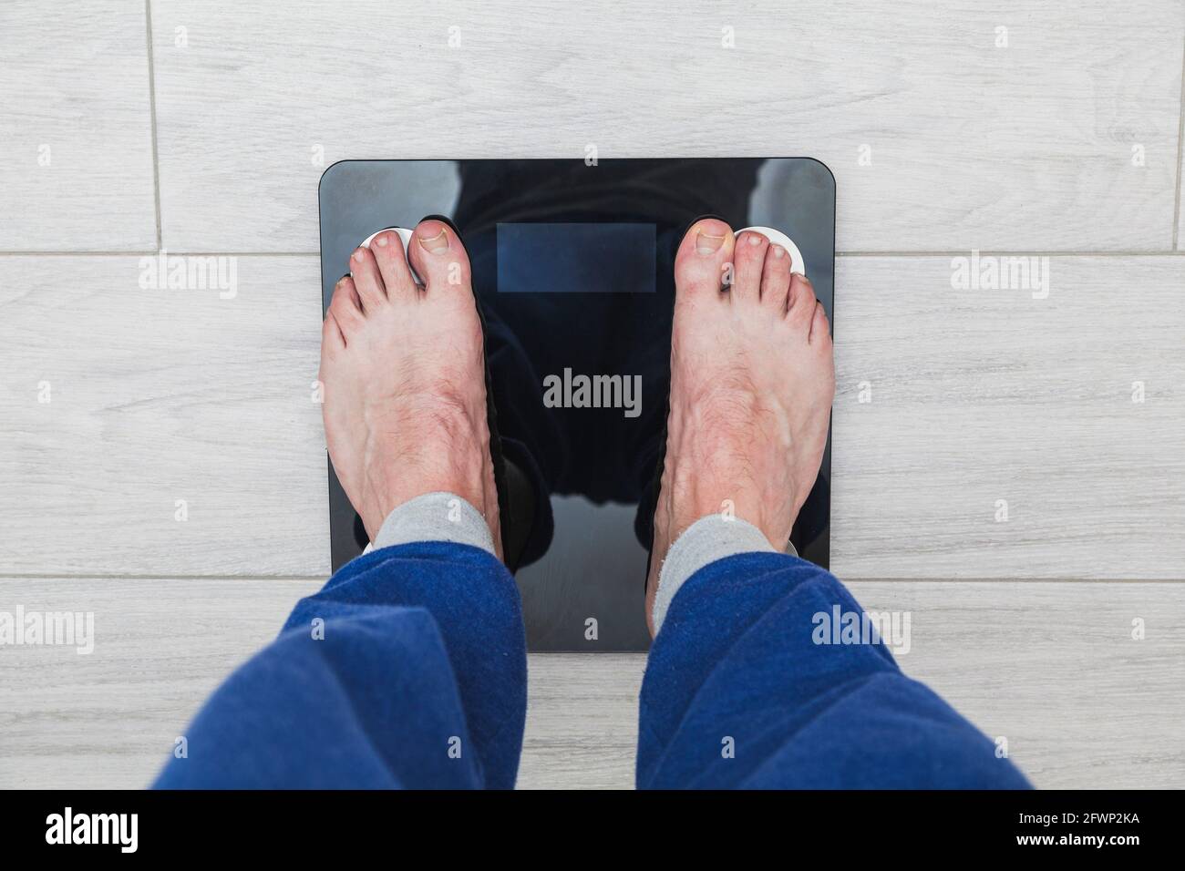 Los pies de un hombre irreconocible encima de una balanza electrónica donde  el peso no es visible. El hombre está usando pantalones de pijama azules y  el suelo es Fotografía de stock -