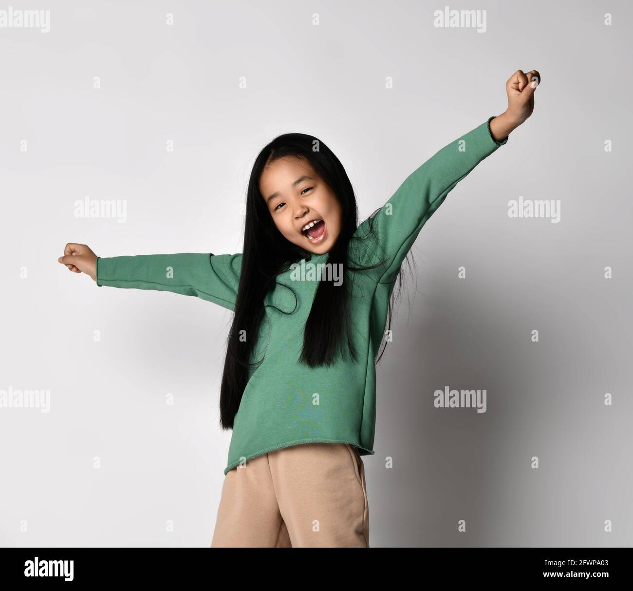 Feliz gritando, cantando en voz alta niña asiática en verde hoodie y pantalones de pie con los brazos hacia arriba y estirado Foto de stock