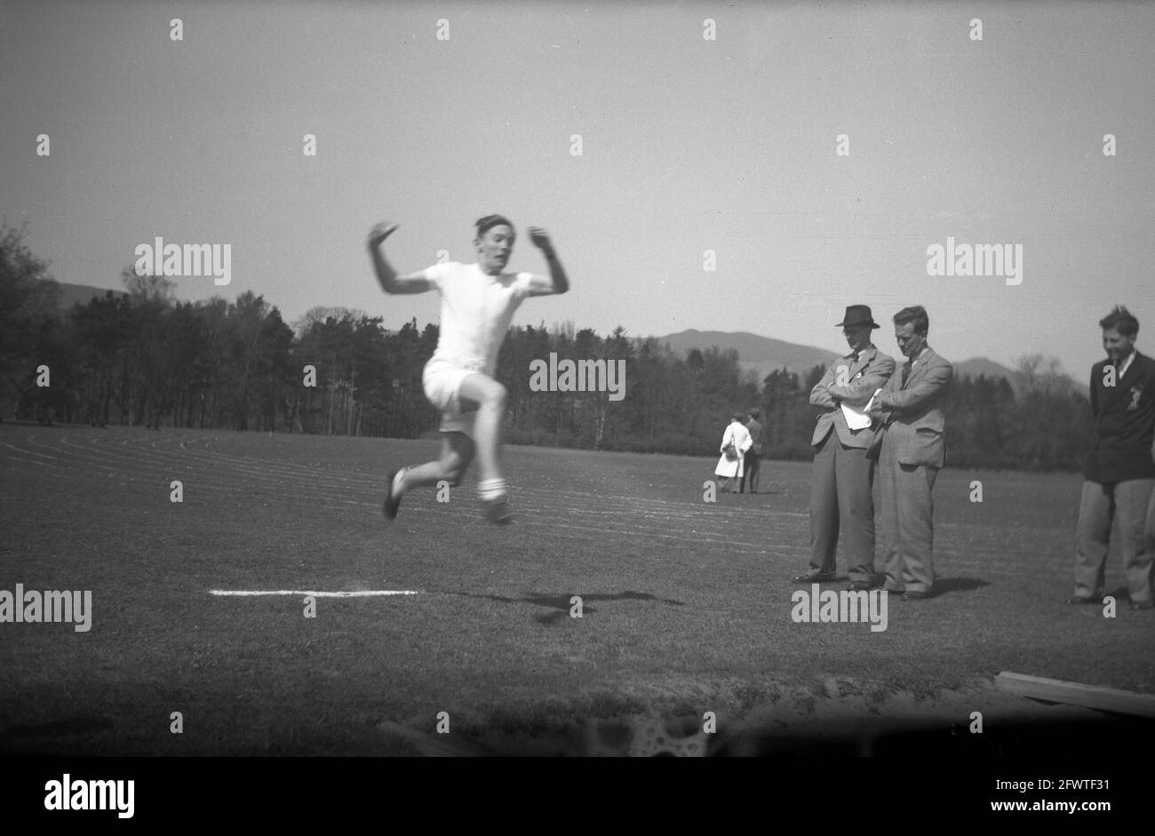 1951, fuera un gran campo de deportes y visto por maestros en la ropa formal la época, uno con un sombrero y compañero de escuela, un joven hombre