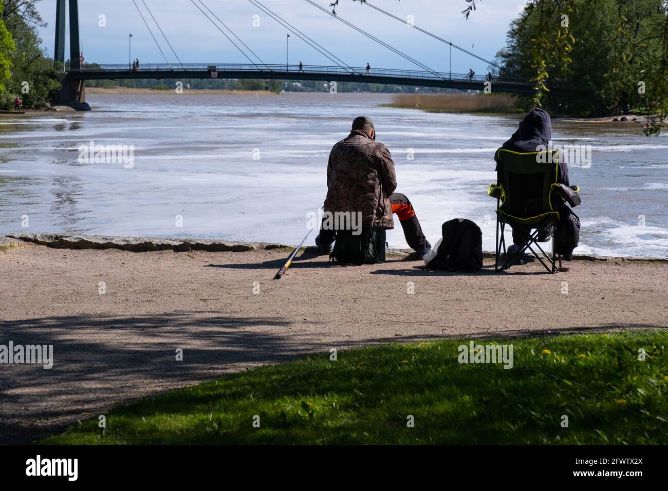 Helsinki / FINLANDIA - 22 DE MAYO de 2021: Dos hombres sentados en la pesca de la costa. Arroyo de agua que conduce hacia un puente peatonal. Foto de stock