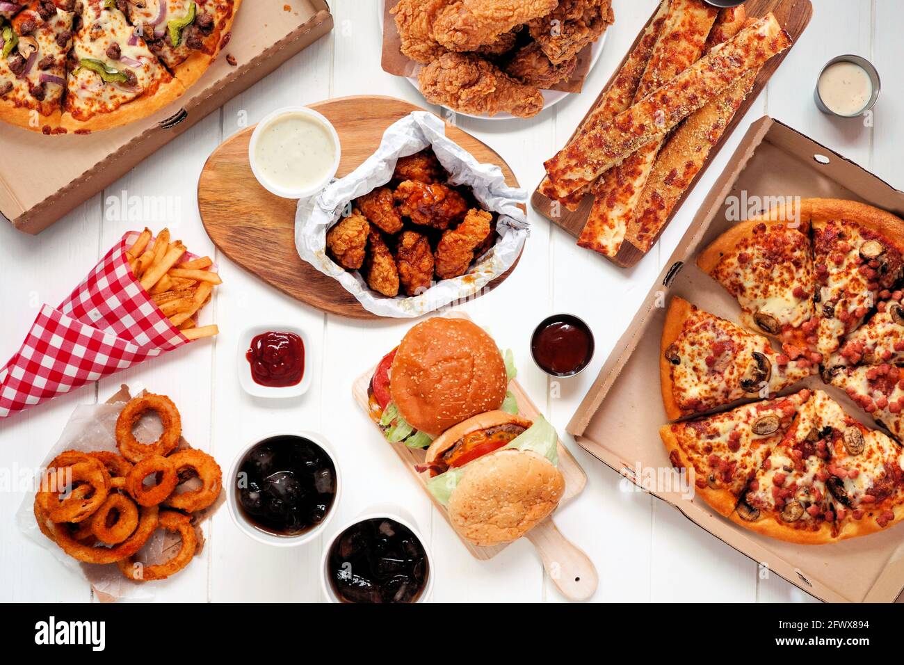 Escena de mesa tipo buffet de comida para llevar o de reparto. Pizza, hamburguesas, pollo frito y sus costados. Vista superior sobre un fondo de madera blanco. Foto de stock