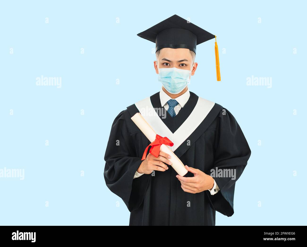 graduación masculina joven usando máscara de la cara y sosteniendo su universidad certificado durante la pandemia de coronavirus Foto de stock