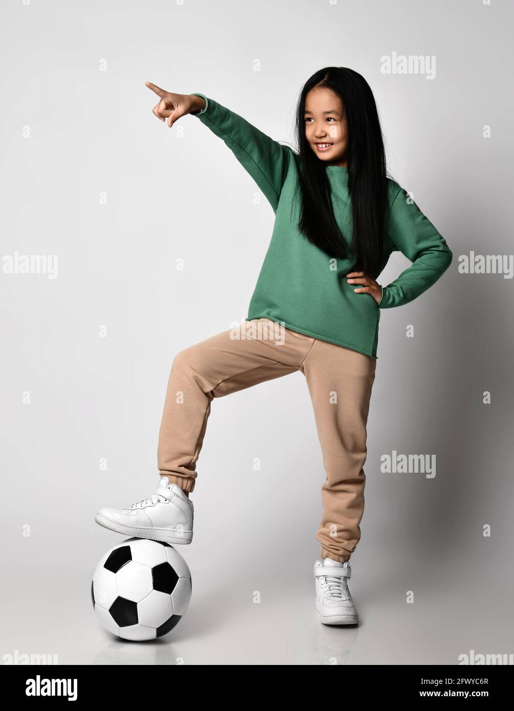 Niña asiática de ropa deportiva verde y zapatillas de deporte blancas se  coloca con balón de fútbol bajo su pie, apuntando con el dedo Fotografía de  stock - Alamy