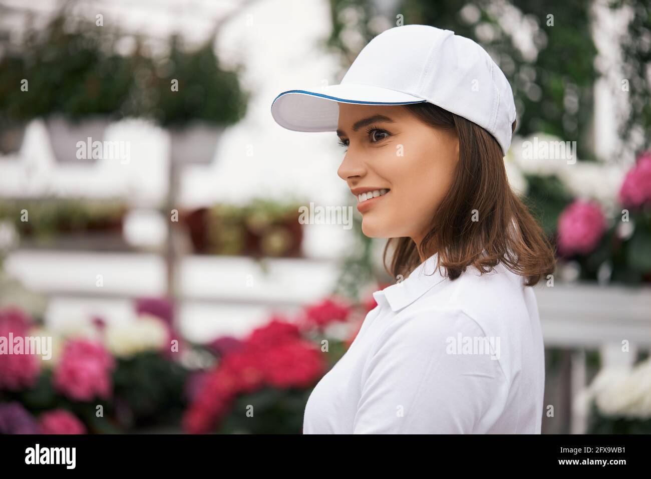 Mujer agradable en gorra blanca camiseta en el invernadero con varias flores de colores creciendo en ollas. Concepto de y agricultura Fotografía de stock -