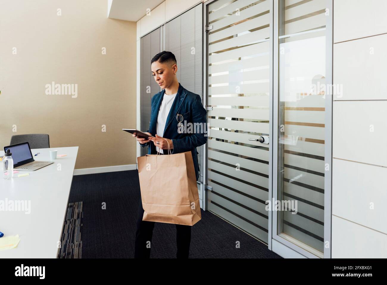 Mujer de negocios que utiliza una tableta digital mientras está de pie con una bolsa de papel en la oficina Foto de stock