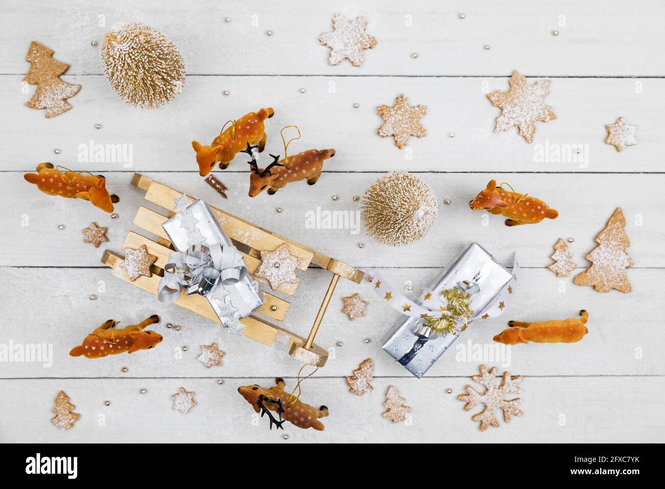 Regalos de Navidad y trineo de juguete rodeado de ciervos de juguete con galletas caseras sobre fondo blanco de madera Foto de stock