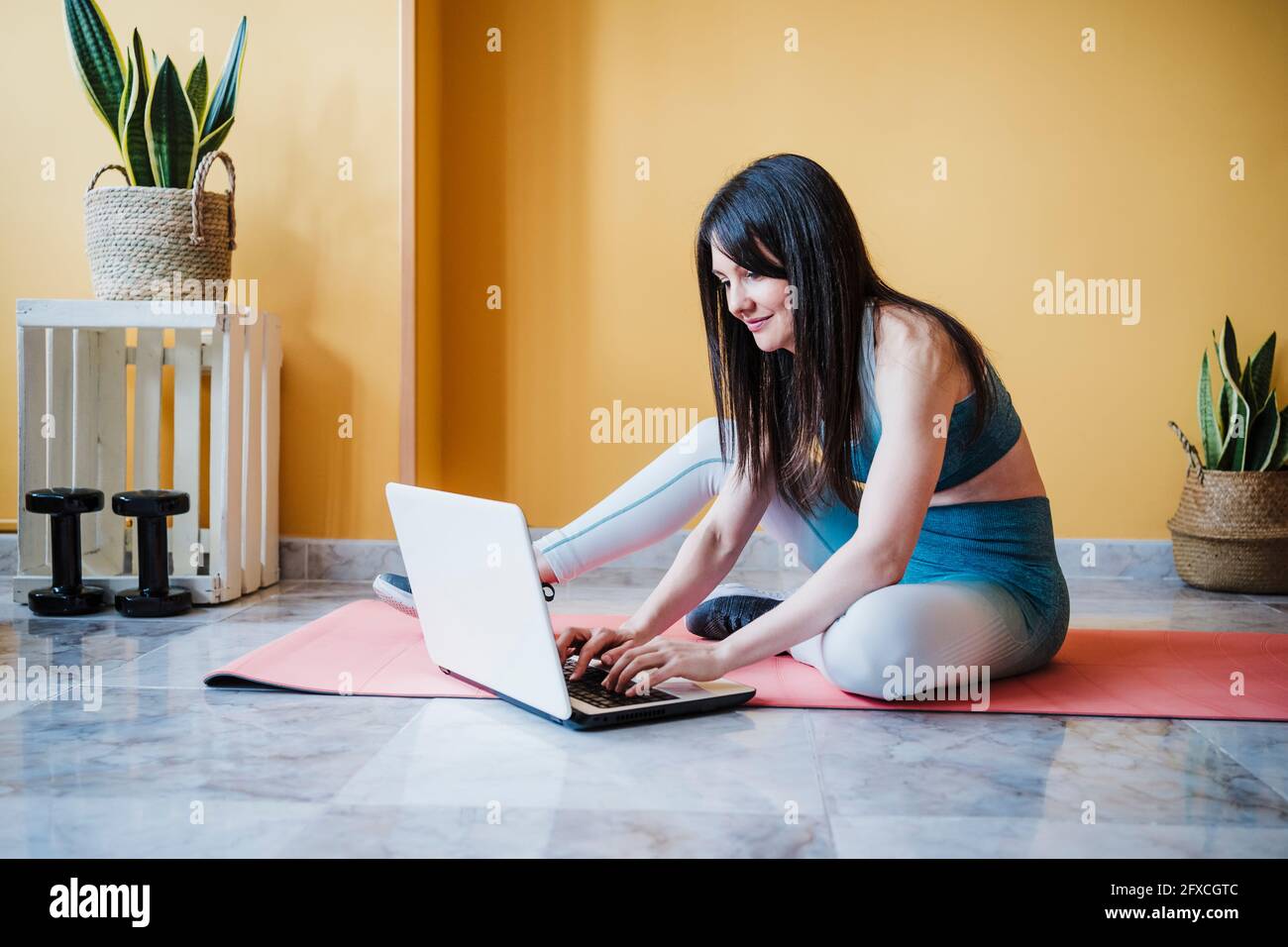 Mujer deportiva que utiliza el portátil mientras se sienta en la colchoneta de ejercicio en casa Foto de stock