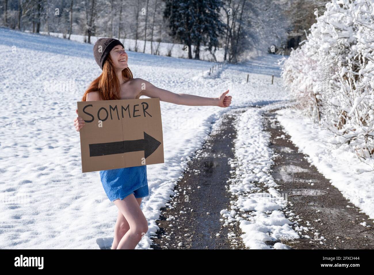 Mujer sonriente con letrero de tablero haciendo senderismo en la carretera cubierta de nieve Foto de stock