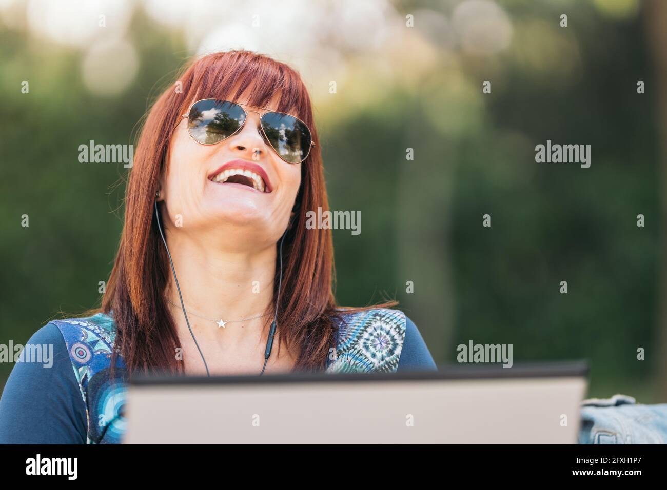 Mujer de pelo rojo sentada en un banco del parque con un ordenador portátil y riendo en voz alta en un día soleado. Primer plano medio. Foto de stock