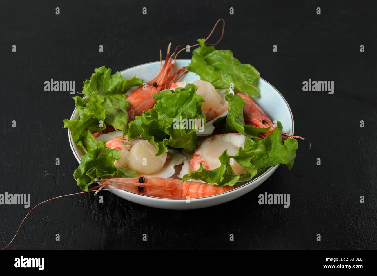 Vieiras crudas naturales con camarón hervido en el fregadero en un tazón de cerámica con hojas de lechuga sobre un fondo oscuro. Enfoque selectivo. Foto de stock