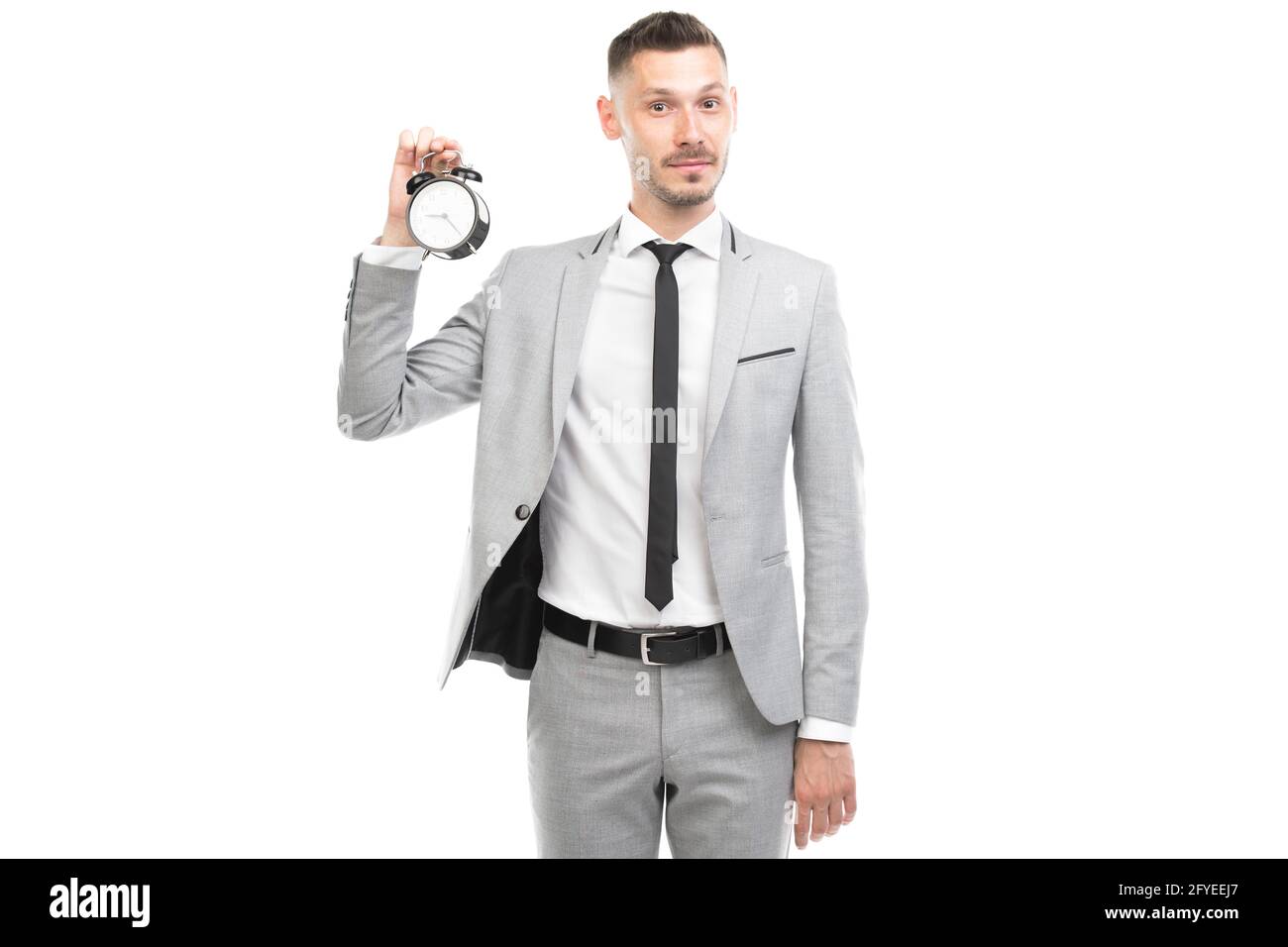 Retrato medio horizontal de un hombre caucásico joven adulto con traje gris que sostiene el reloj despertador, concepto de fecha límite, fondo blanco Foto de stock