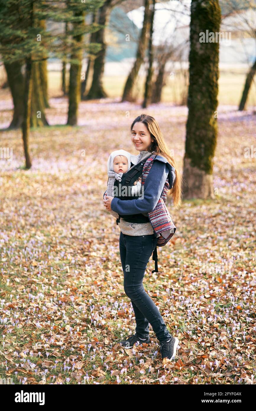 Mamá en un chaleco y jeans sostiene a un bebé en mochila de canguro en un  sombrero blanco con orejas de pie en el bosque Fotografía de stock - Alamy