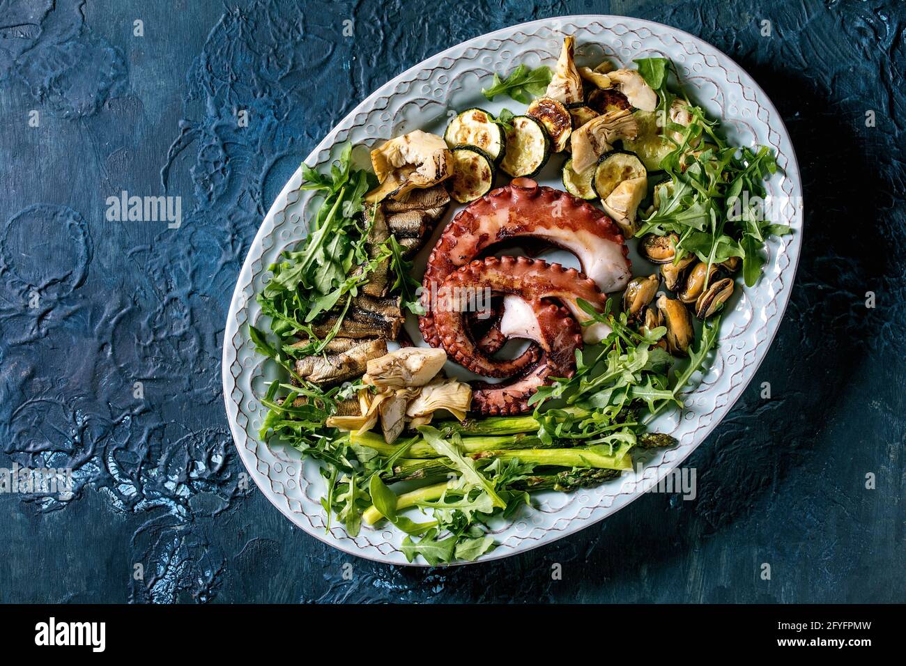 Ensalada de marisco. Se cobaron tentáculos de pulpo, sardinas y mejillones a la parrilla sobre un plato de cerámica azul con ensalada de rúcula, calabacín y espárragos sobre t azul Foto de stock