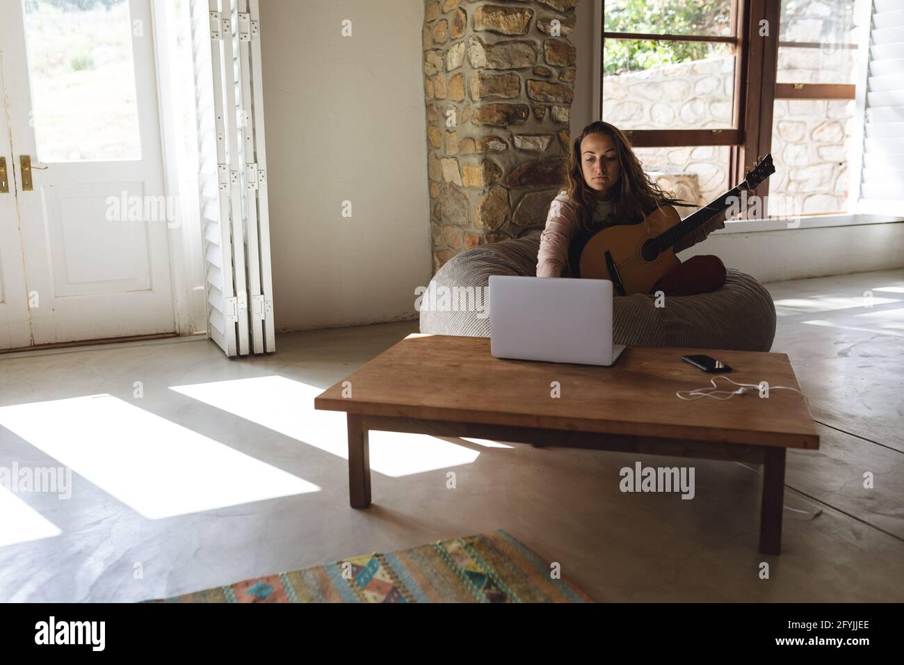 Mujer caucásica sentada en un bolso tocando guitarra acústica usando un ordenador  portátil en la soleada cabaña sala de estar Fotografía de stock - Alamy