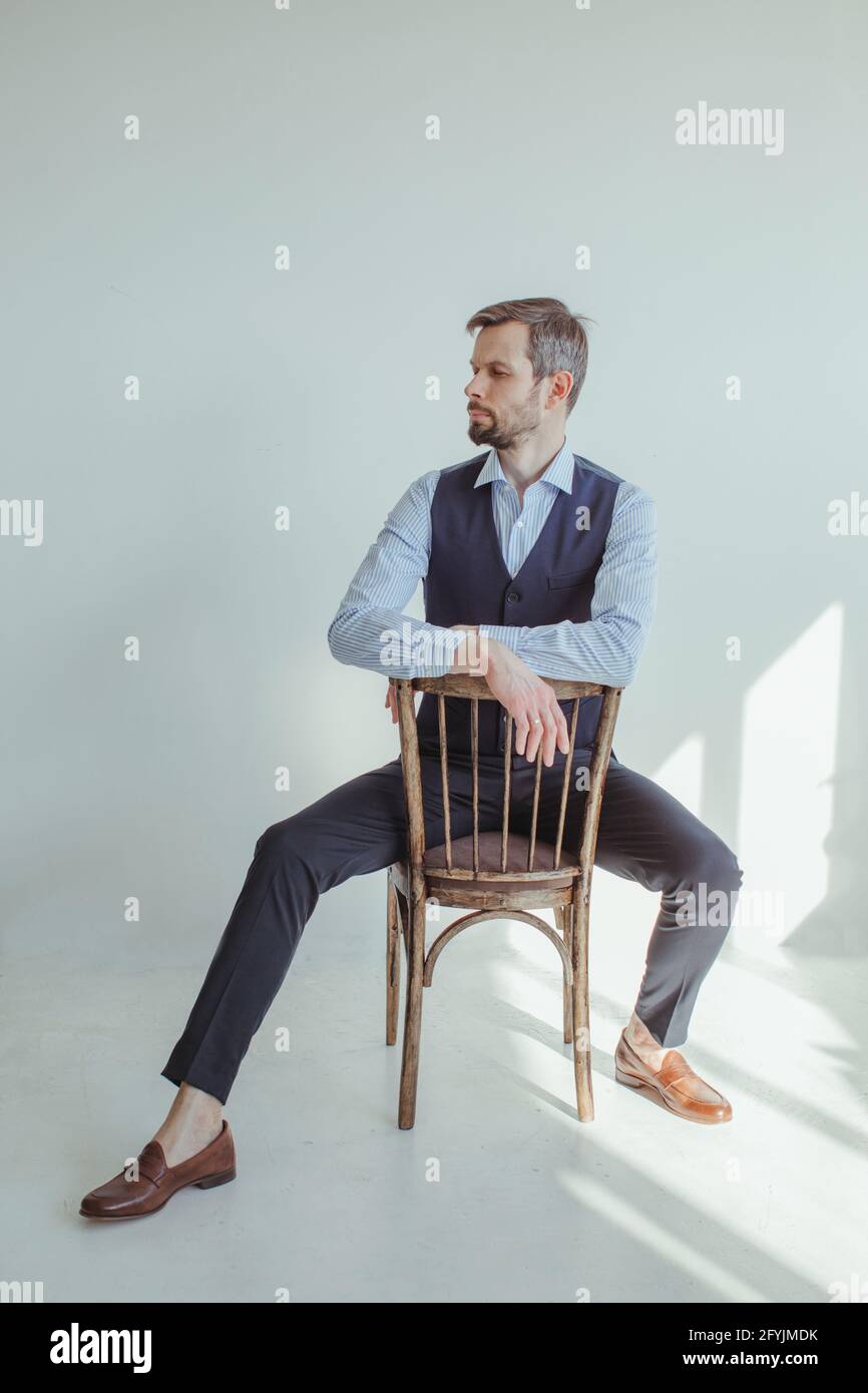 Retrato de un hombre en una camisa y chaleco sentado y apoyado en la parte posterior de una silla bajo la luz del sol Foto de stock