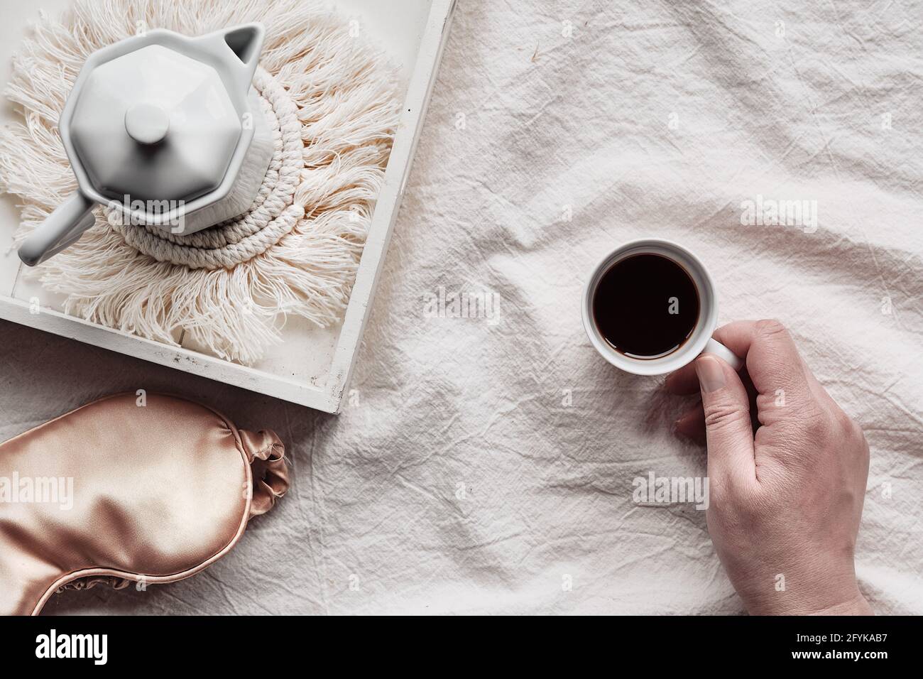 Espresso en la mano, bandeja con cafetera espresso de cerámica blanca en la moderna alfombrilla macrame. Máscara para dormir sobre sábanas arrugadas. Mano femenina con taza de Foto de stock