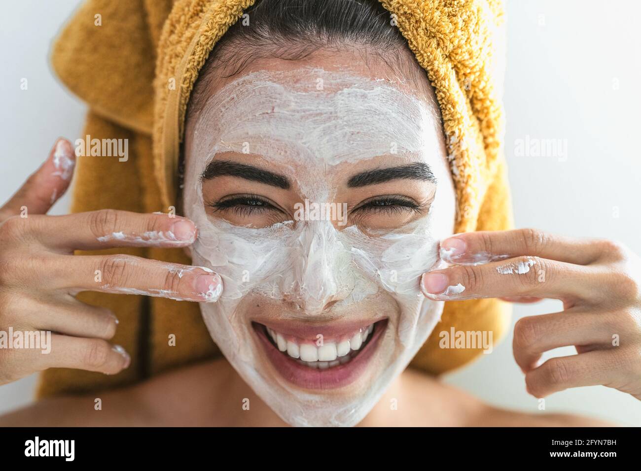 Mujer joven sonriente aplicando crema en la cara - Feliz chica un día de spa para el cuidado de la piel en casa Foto de stock