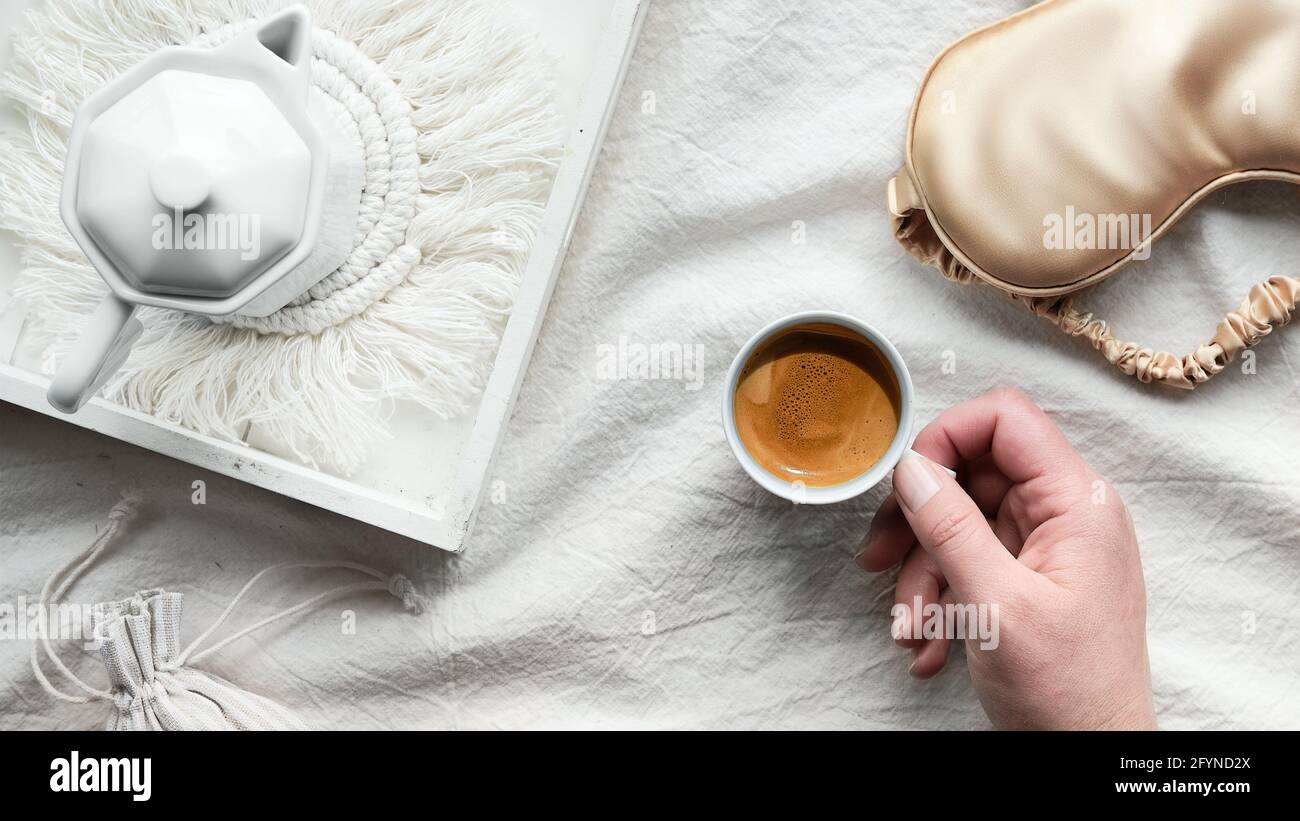 Espresso en la mano, bandeja con cafetera espresso de cerámica blanca en la moderna alfombrilla macrame. Máscara para dormir sobre sábanas arrugadas. Mano femenina con taza de Foto de stock