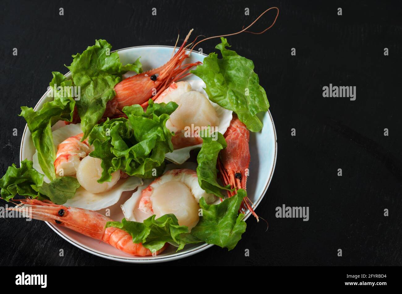 Vieiras crudas naturales con camarón hervido en el fregadero en un tazón de cerámica con hojas de lechuga sobre un fondo oscuro. Enfoque selectivo. Foto de stock