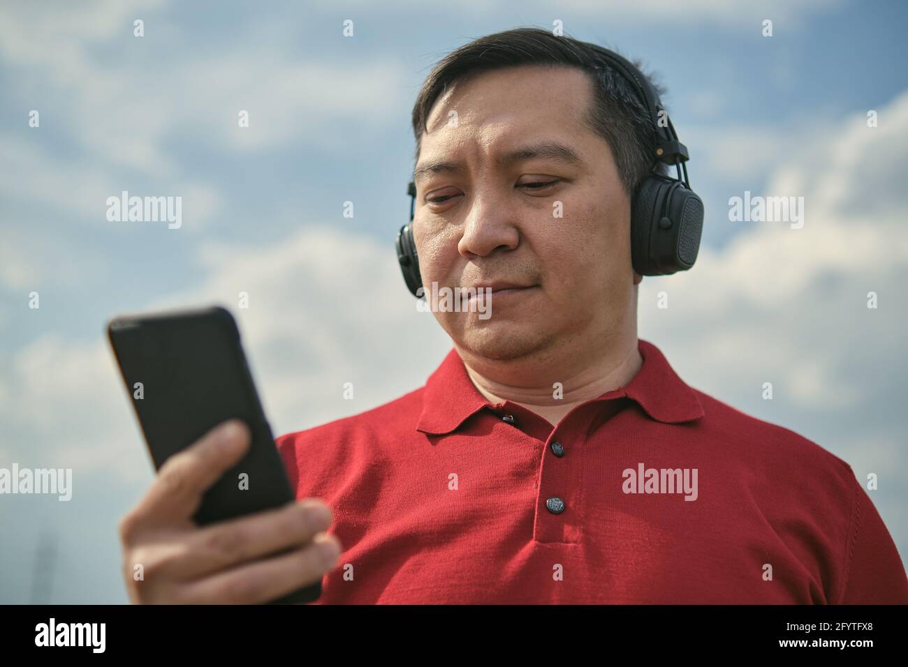 Hombre asiático de mediana edad con auriculares al aire libre escuchando música sobre el fondo del cielo, teléfono móvil en la mano Foto de stock
