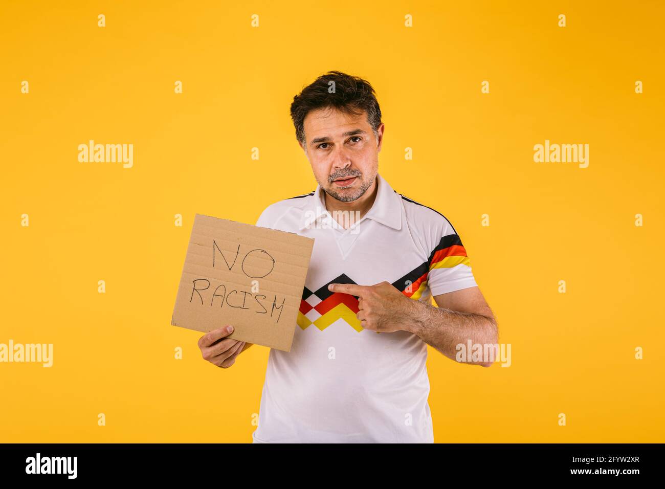 Un aficionado al fútbol que lleva una camiseta blanca con rayas negras, rojas y amarillas, tiene un cartel que dice 'No racismo' Foto de stock