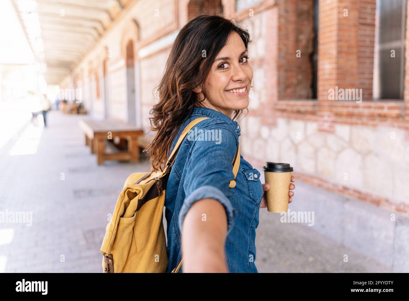 Mujer sonriente con chaqueta vaquera y mochila amarilla con comida para  llevar café mirando la cámara con el gesto de seguirme en la calle cerca  del edificio Fotografía de stock - Alamy