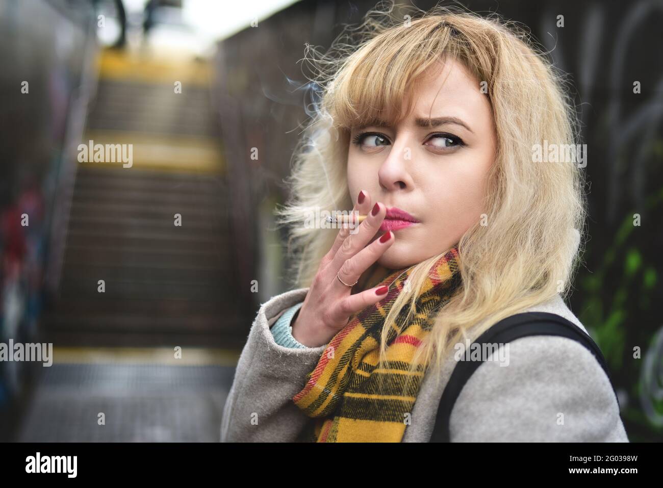 Mujer rubia joven seria que fuma un cigarrillo y mira a un lado en el pasaje subterráneo Foto de stock