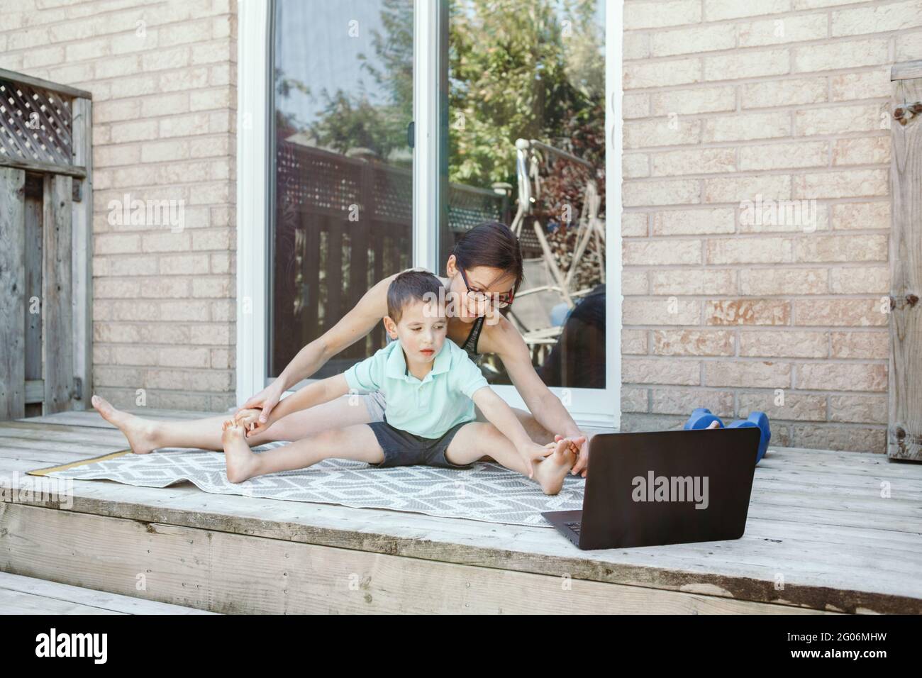 Actividad deportiva familiar. Madre caucásica de mediana edad con hijo pequeño haciendo a distancia ejercicio de fitness en línea en el patio trasero de la casa con ordenador portátil. Padre Foto de stock