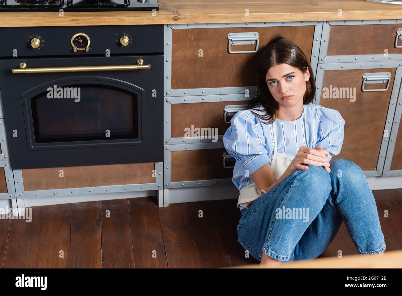 mujer adulta joven cansada sentada en el suelo cerca del horno y en la cocina moderna Foto de stock