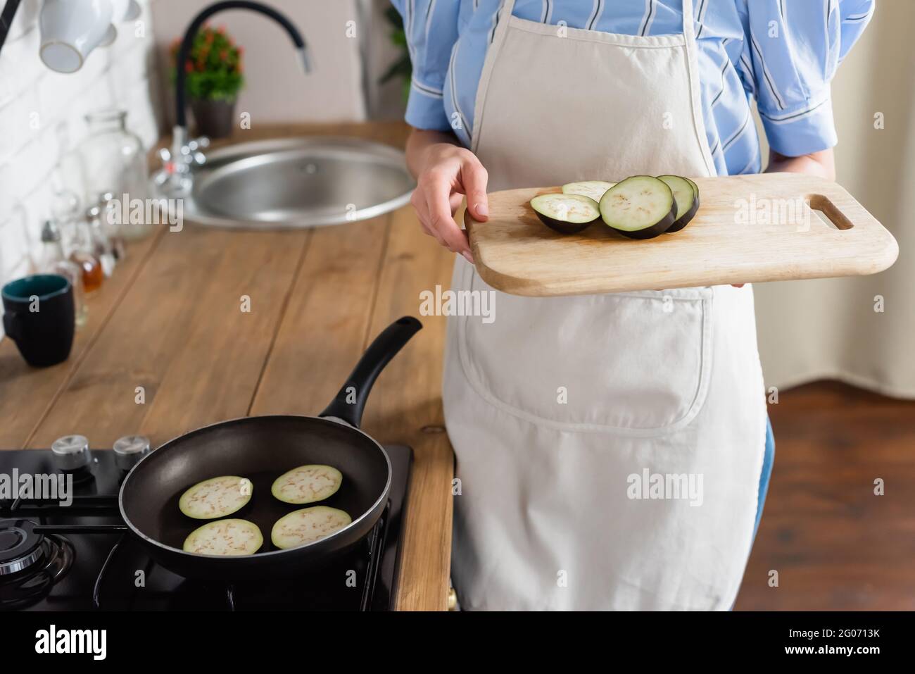 vista recortada de una mujer adulta joven sosteniendo rebanadas de berenjena en una tabla de cortar cerca de una sartén en la cocina Foto de stock