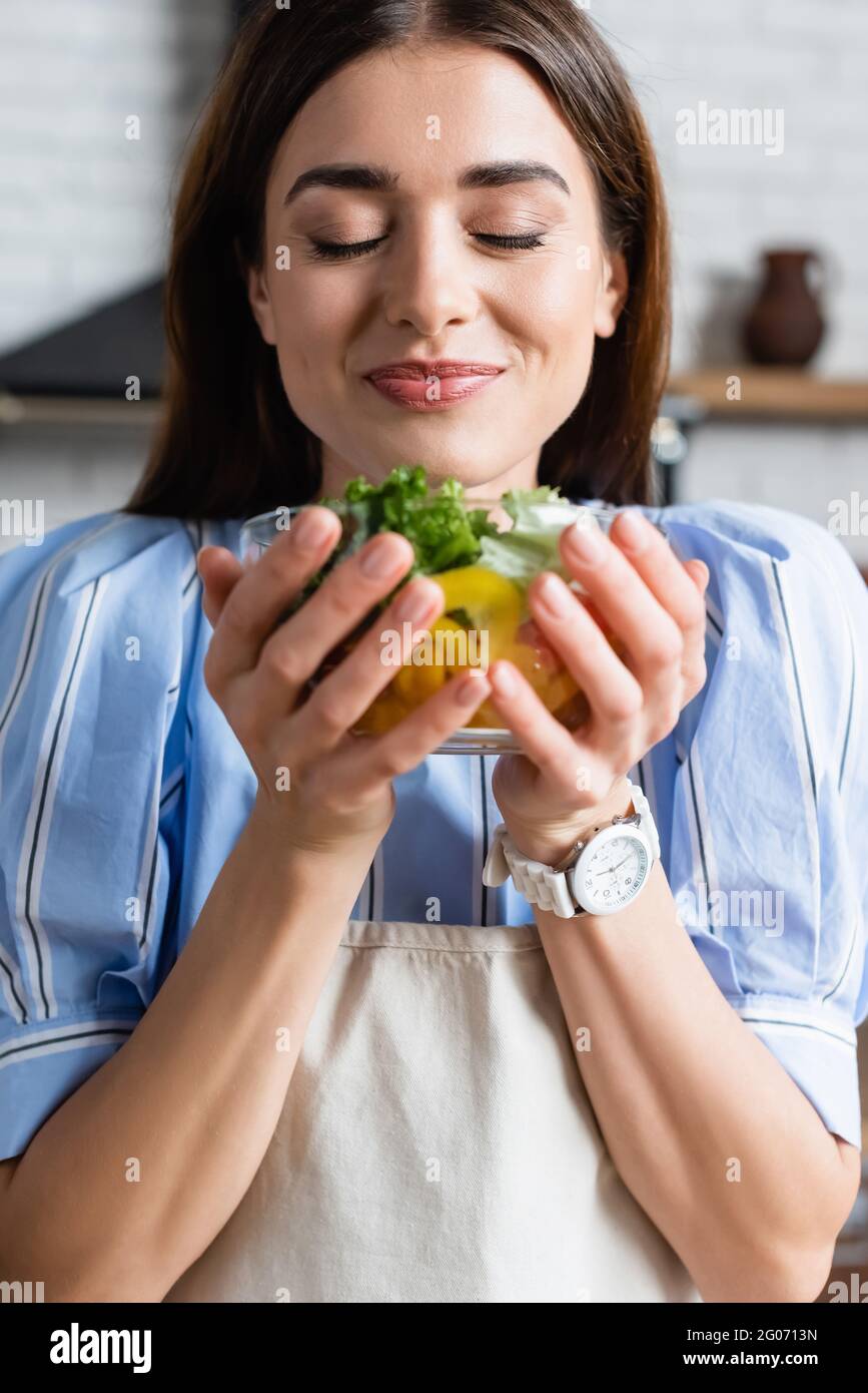 sonriente joven adulta disfrutando de un aroma a ensalada de verduras frescas con ojos cerrados en la cocina Foto de stock