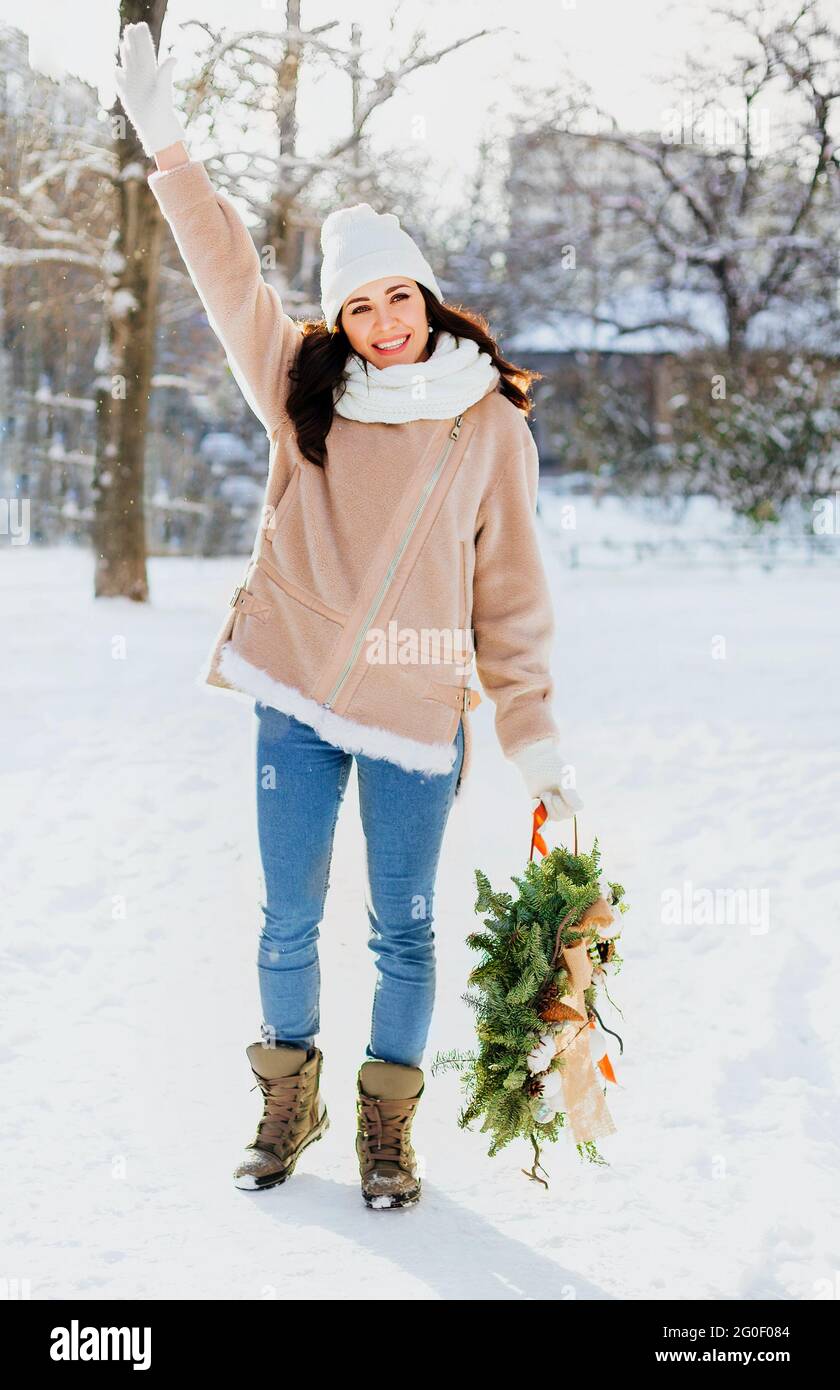Feliz mujer en ropa exterior con corona de Navidad decorada y de pie en la nieve en el soleado día de invierno Foto de stock