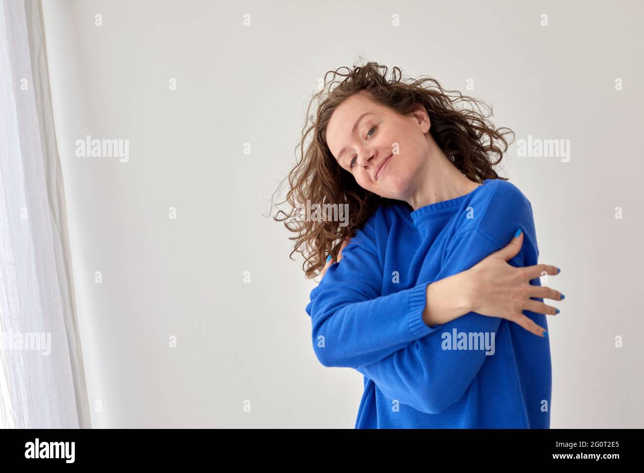 Abrazo de la joven confiada feliz contra fondo blanco de la pared. Vista lateral del contenido femenino con un moderno suéter azul sobre fondo gris Foto de stock