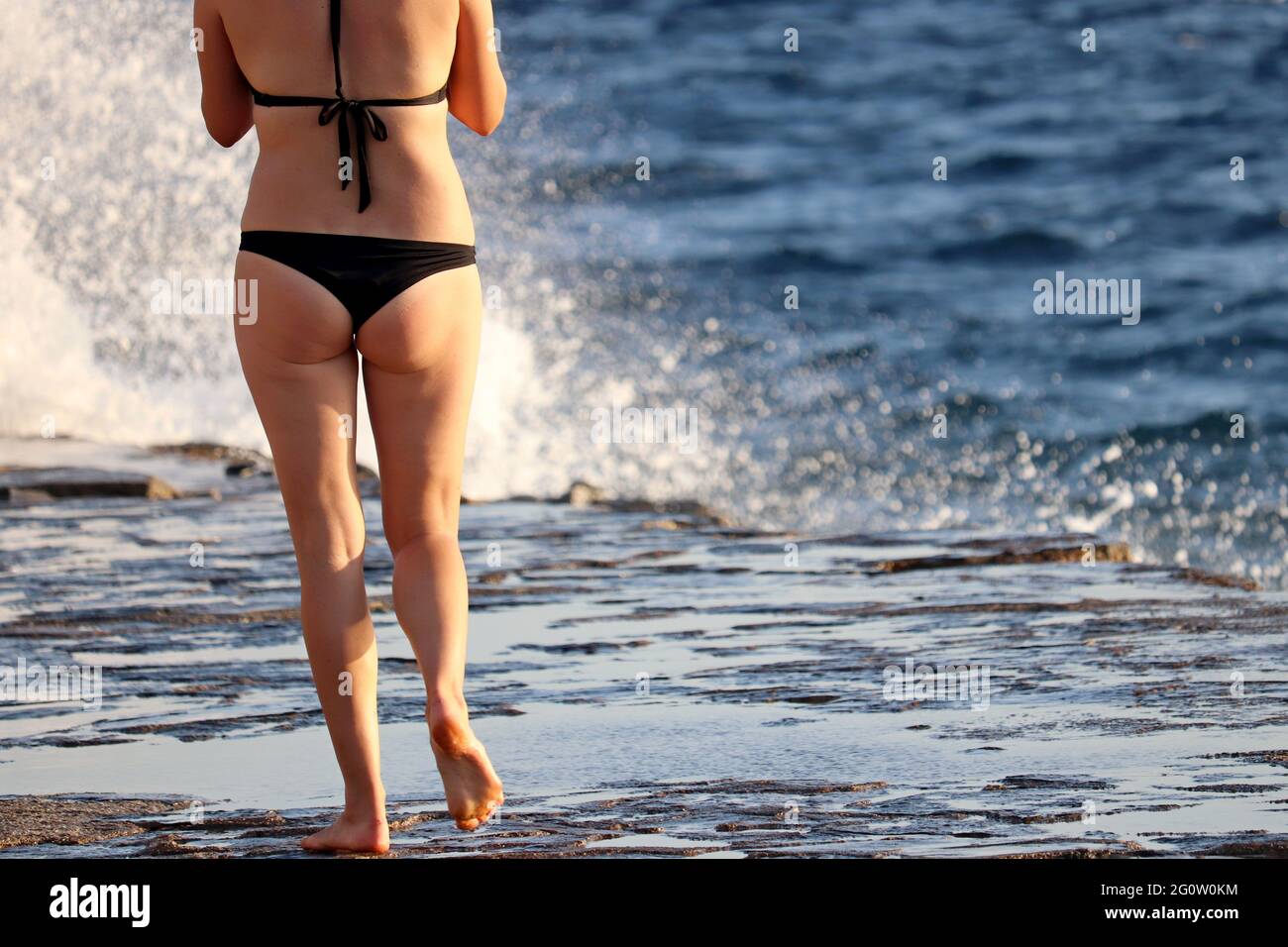 Vacaciones en la playa, mujer en bikini negro caminando en la costa de  piedra sobre el mar azul y el agua salpican el fondo Fotografía de stock -  Alamy