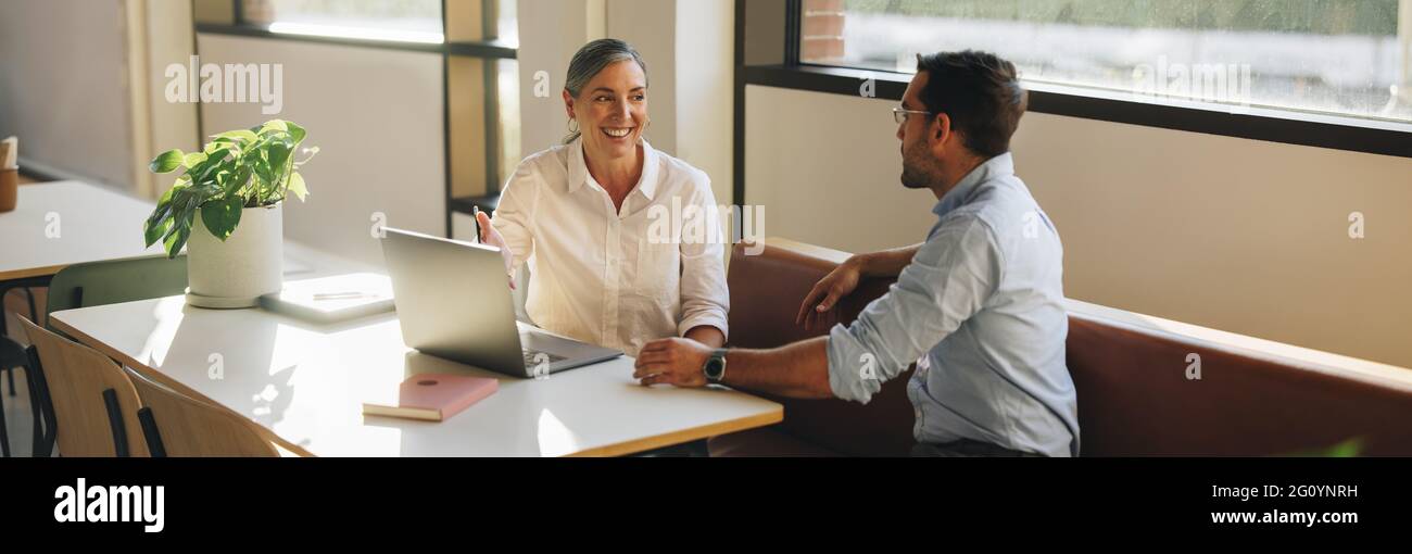 Mujer de negocios que se ve feliz durante una reunión con un compañero en el escritorio de la oficina. Mujer sonriente apuntando al portátil y hablando con su compañero de trabajo en la oficina. Foto de stock