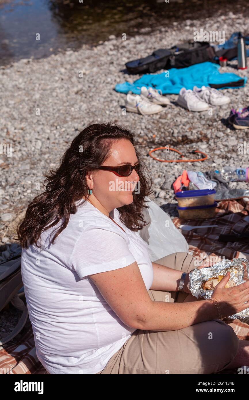 La mujer joven toma un descanso mientras hace senderismo y tiene un pollo asado en su mano Foto de stock