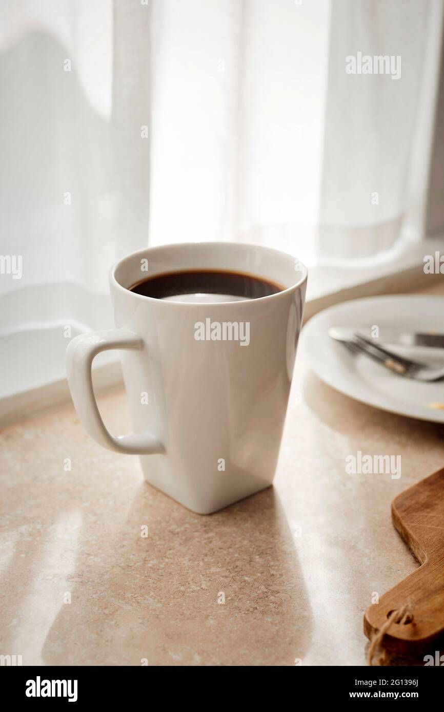 Una taza blanca de café sentado en una encimera de una cocina con sol. Foto de stock