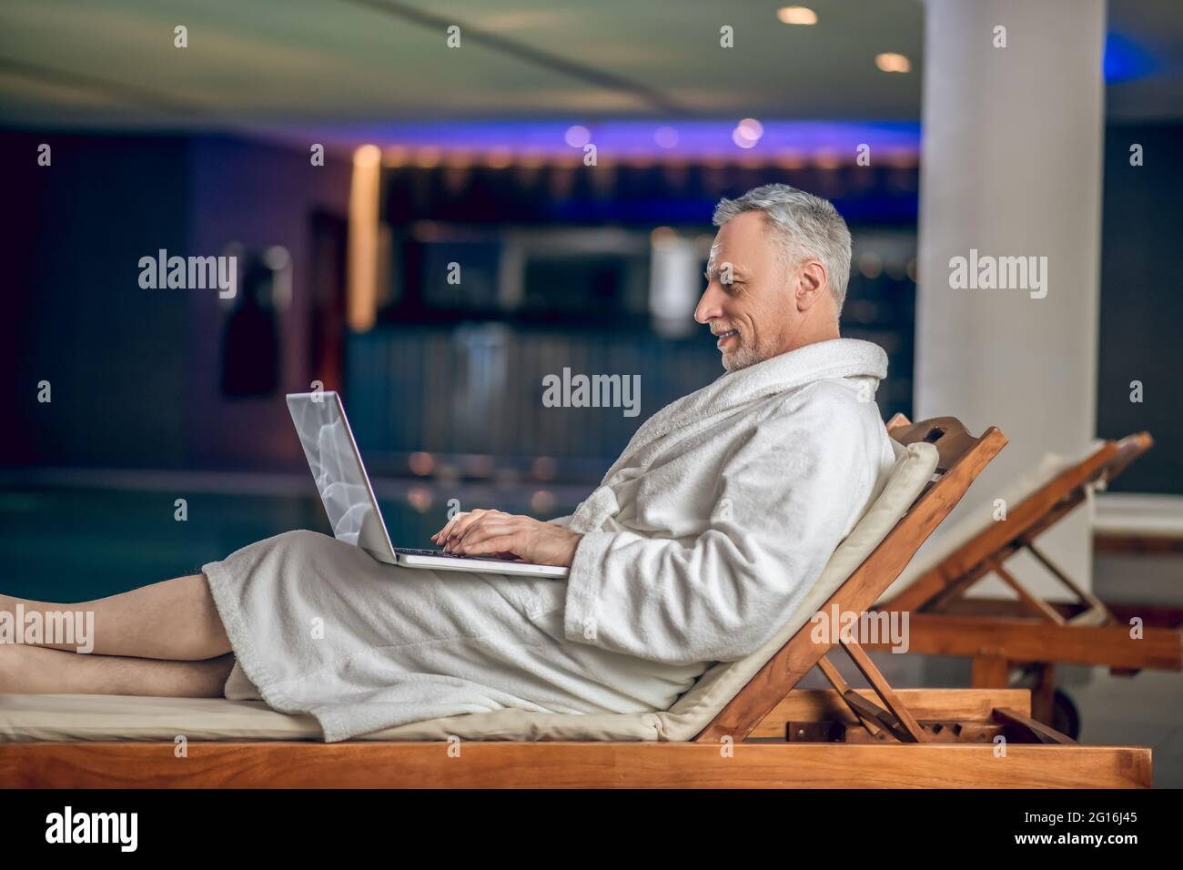 Un hombre barbudo de pelo gris en una bata de baño blanca que pasa tiempo en Internet Foto de stock