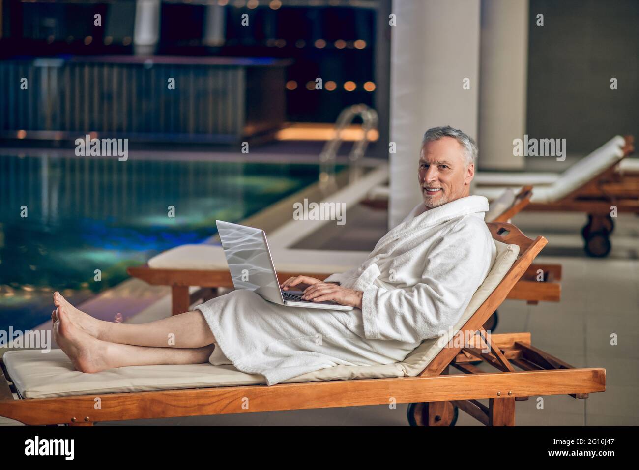 Un hombre barbudo de pelo gris en una bata de baño blanca que pasa tiempo en Internet Foto de stock